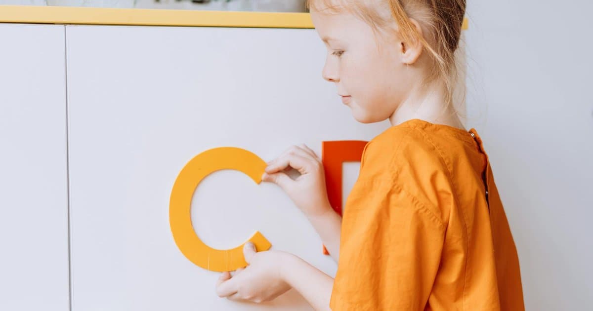 Young girl in orange shirt arranging colorful letters on a surface for alphabet learning practice.