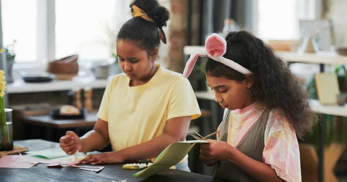 Two girls with bunny ears doing a paper craft project, representing creative gift ideas for young readers.
