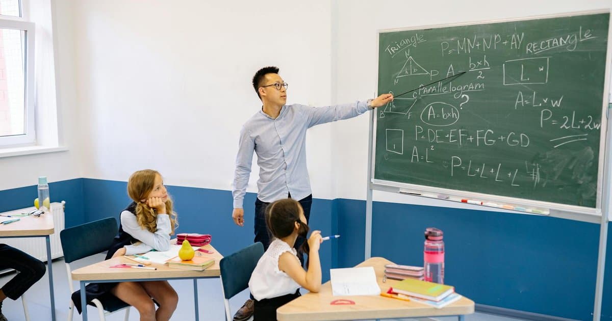 Teacher pointing at a geometry diagram on a chalkboard while students watch in a classroom.