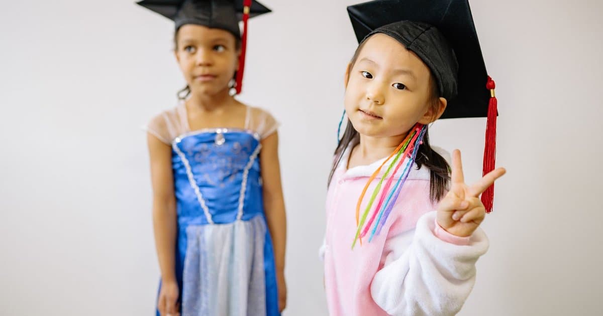 Two young children wearing graduation caps smile together, representing preschool end-of-year celebrations.