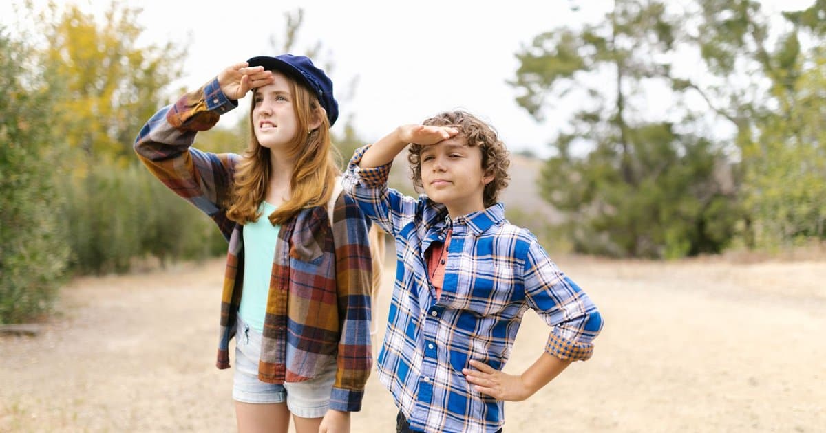Two children in plaid shirts look off into the distance outdoors while searching for clues.