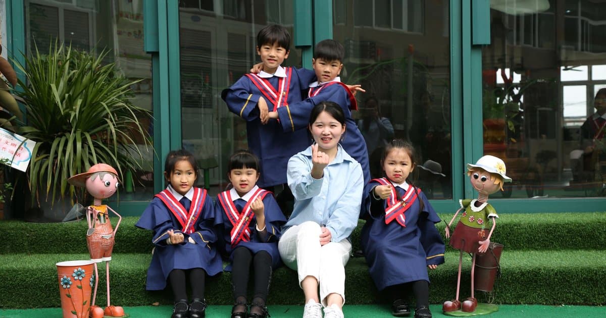 Children in blue graduation caps and gowns pose with a teacher outdoors for a kindergarten graduation celebration.