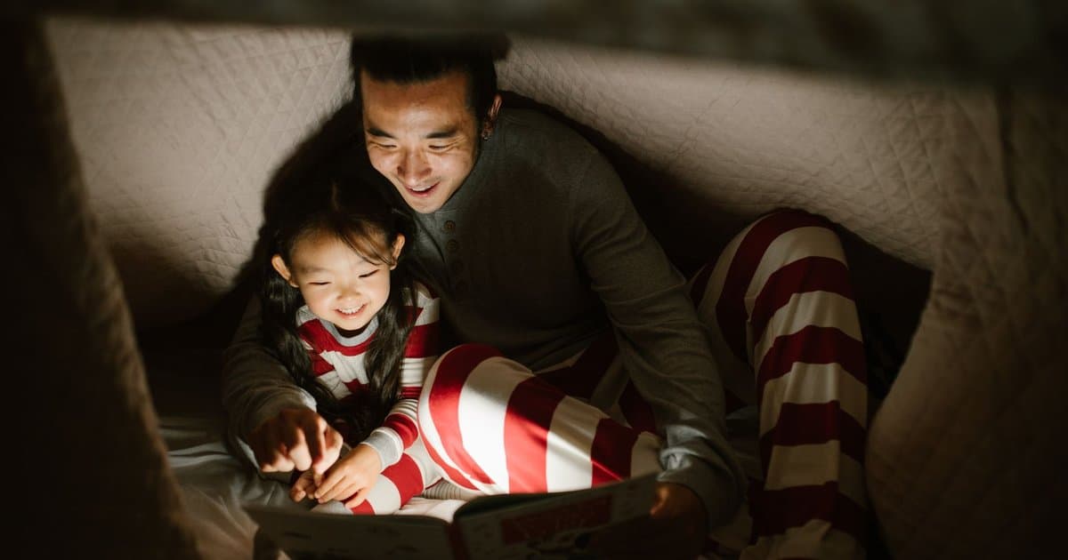 Father and daughter reading together under a blanket fort with a flashlight, sharing a book moment.