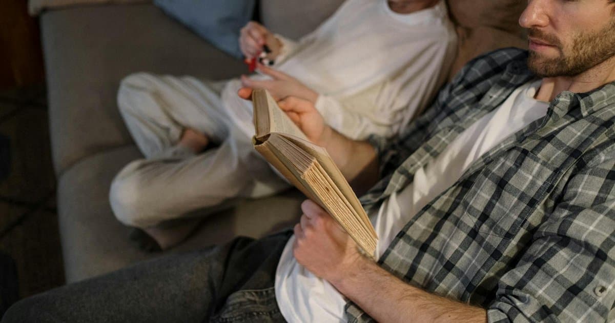 Father reading a book while his son plays nearby on a couch, illustrating meaningful book gift ideas for fathers.
