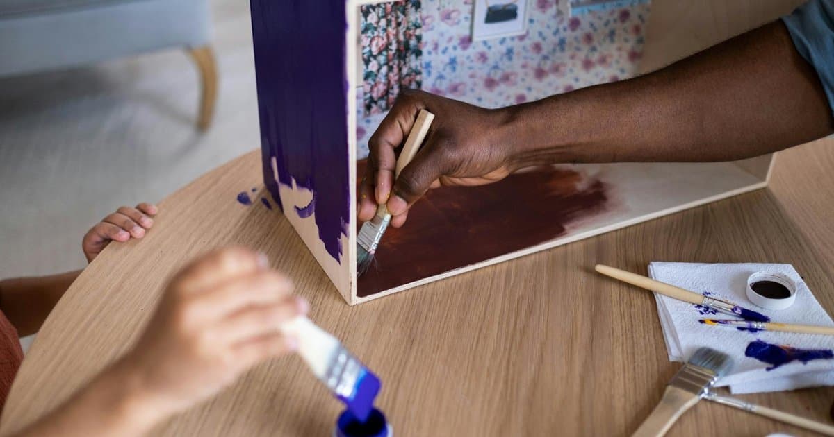 A parent and toddler paint a wooden dollhouse together with colorful brushes, making a handmade gift.