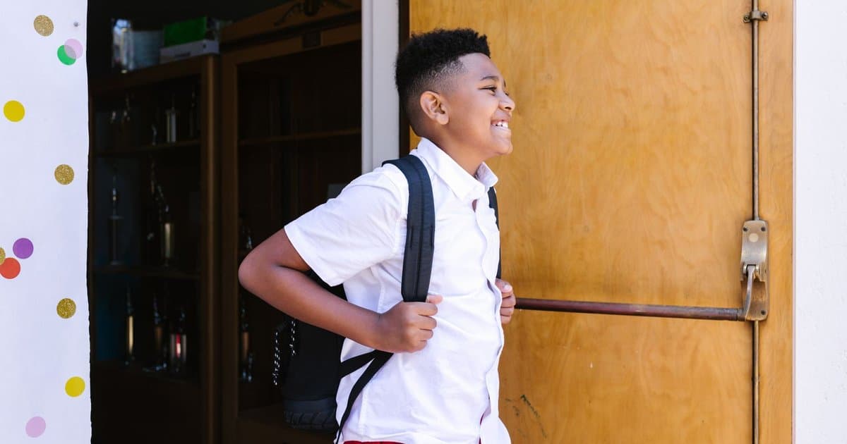 A smiling boy with a backpack entering school, representing the transition to first grade.