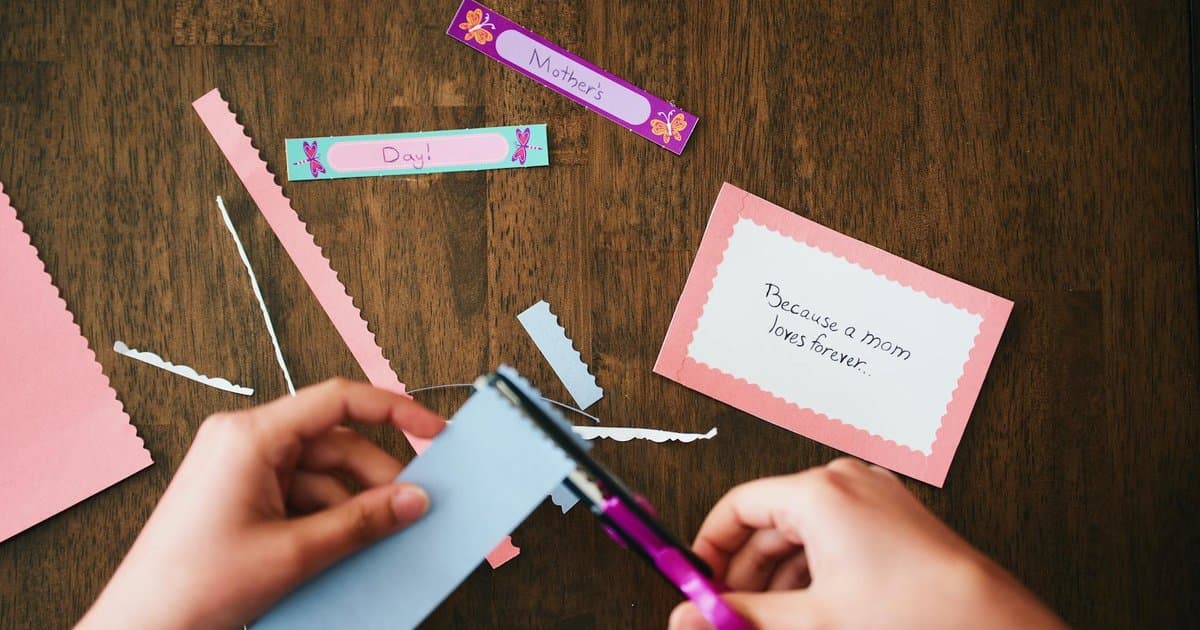 Child making a handmade Mother's Day card with colorful paper and scissors on a table.