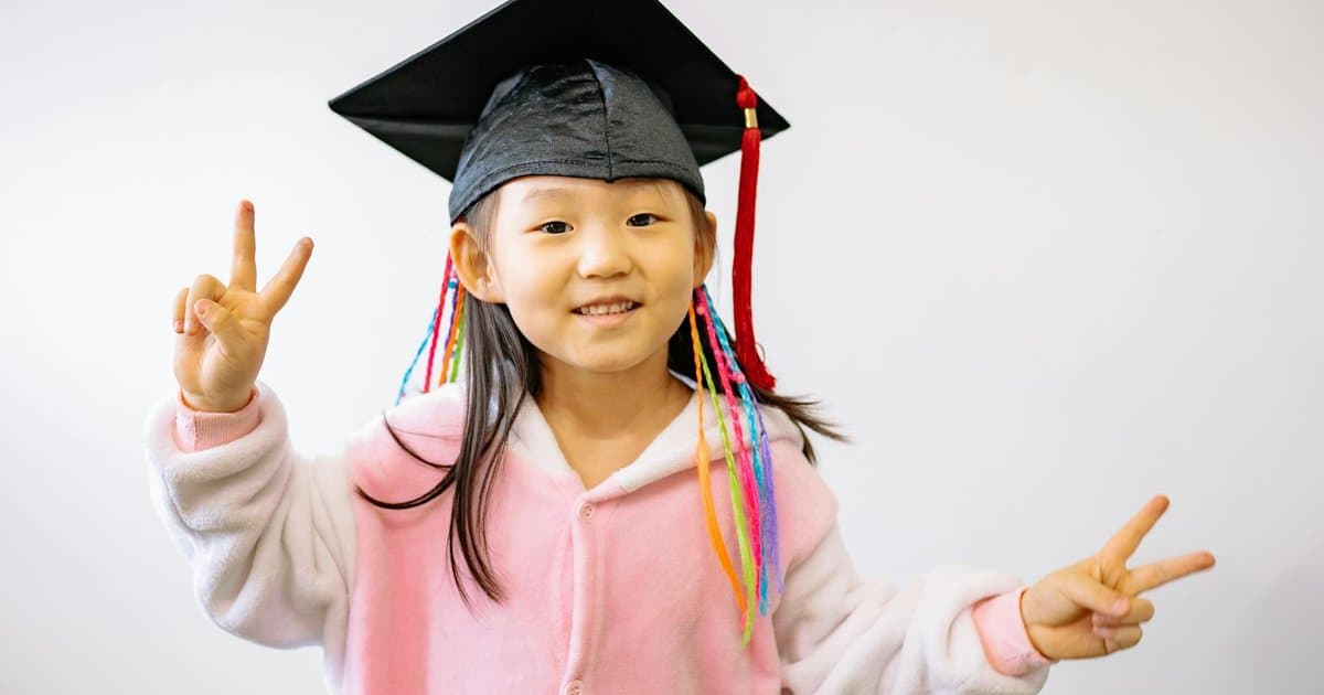 A smiling child wearing a graduation cap and making peace signs, symbolizing kindergarten milestones and gift ideas.