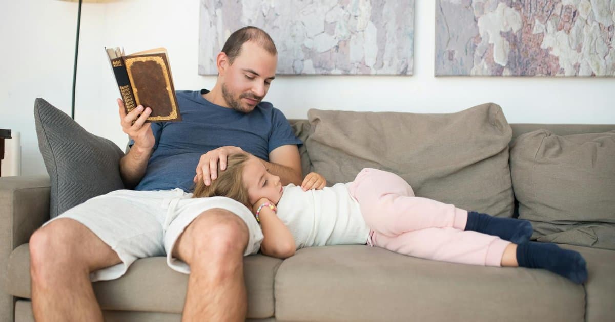 A father reads a book while his daughter sits beside him on a couch at home.