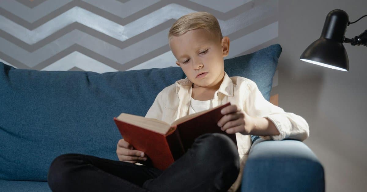 Boy reading on a blue couch under lamp light, illustrating the engagement personalized books create for this age group.