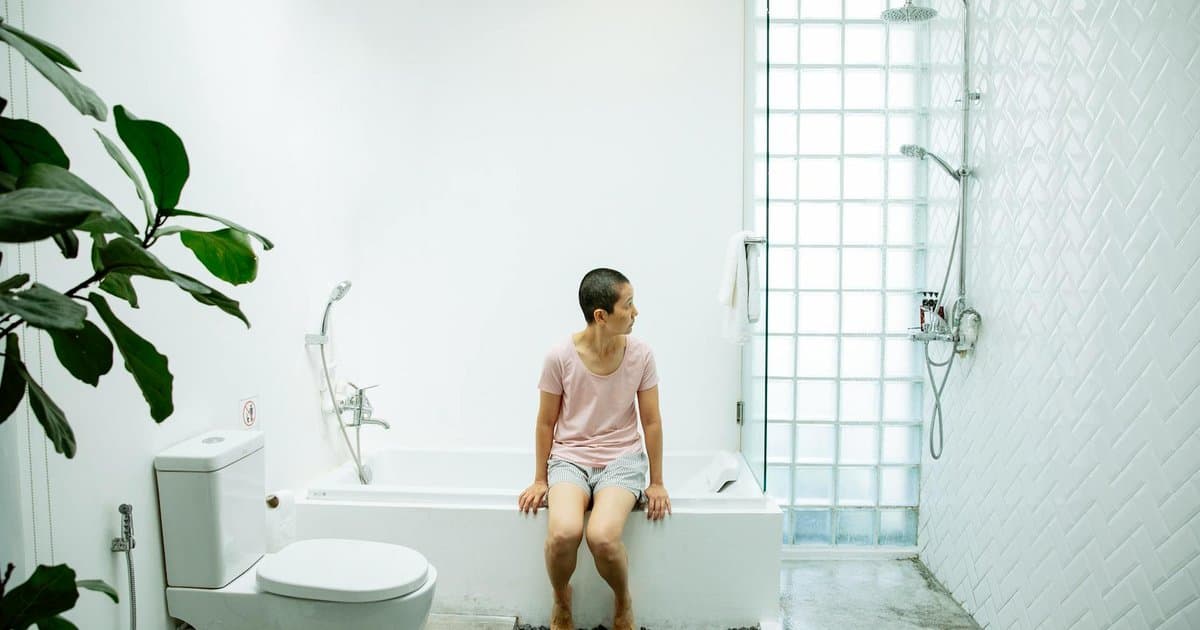 Woman sitting on bathtub in bathroom near toilet, illustrating potty training method comparison.