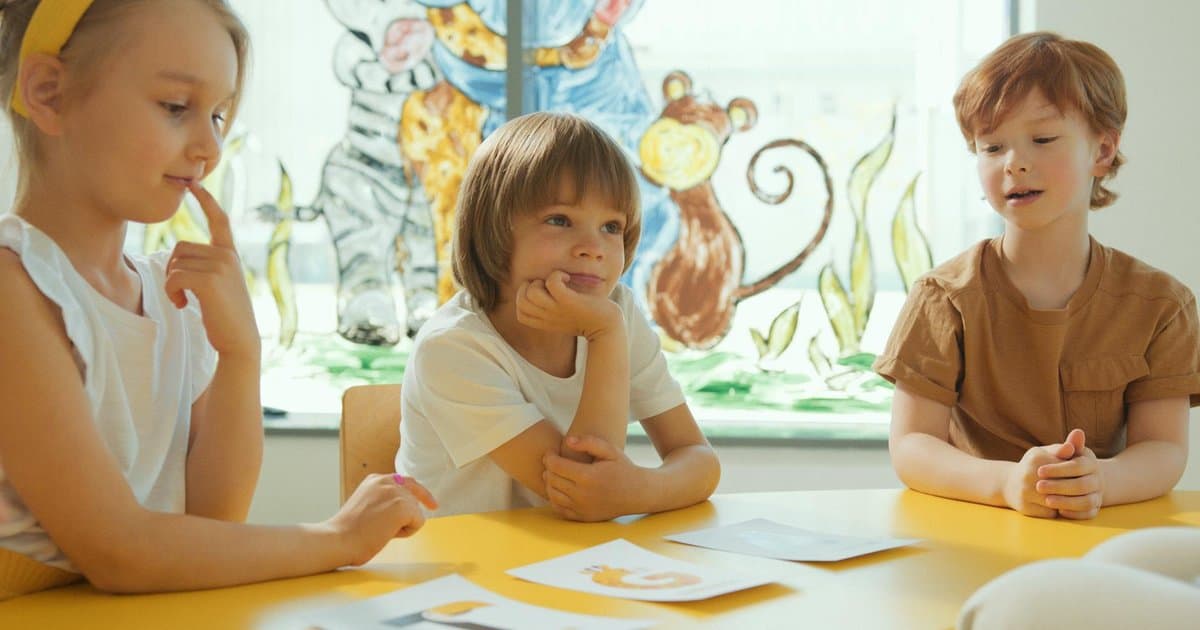 Three children sit at a yellow classroom table working on a learning activity together.