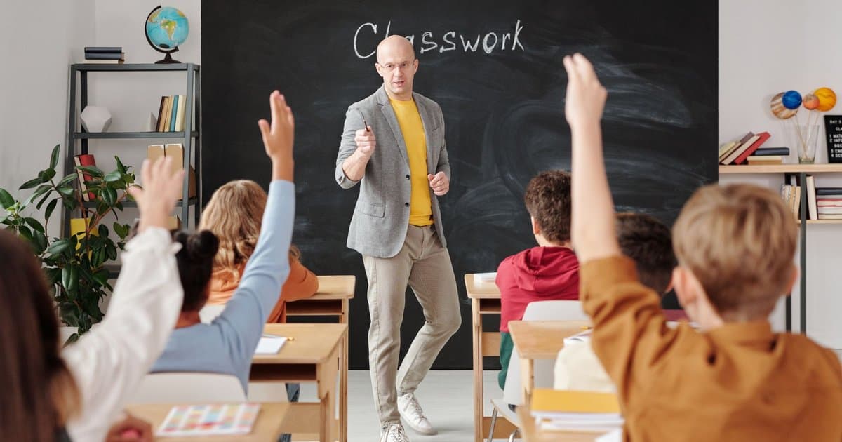 Teacher at desk with students raising hands in classroom, illustrating what teachers value in the classroom.
