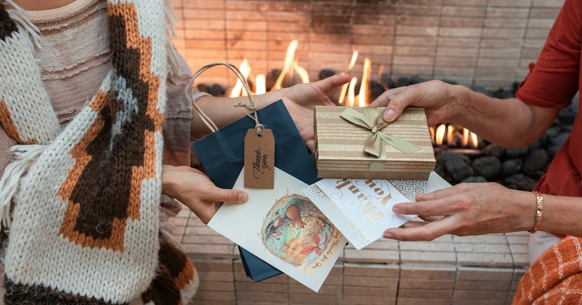 Two people exchanging wrapped gifts by a fireplace with cards and presents nearby, representing thoughtful gift-giving for teachers.
