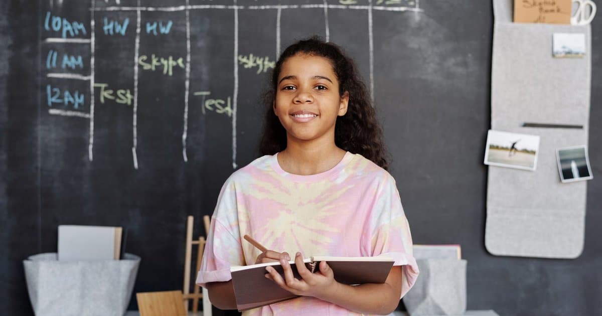 Teen girl holding a notebook in a classroom with a chalkboard timetable behind her, showing a visual schedule tool.