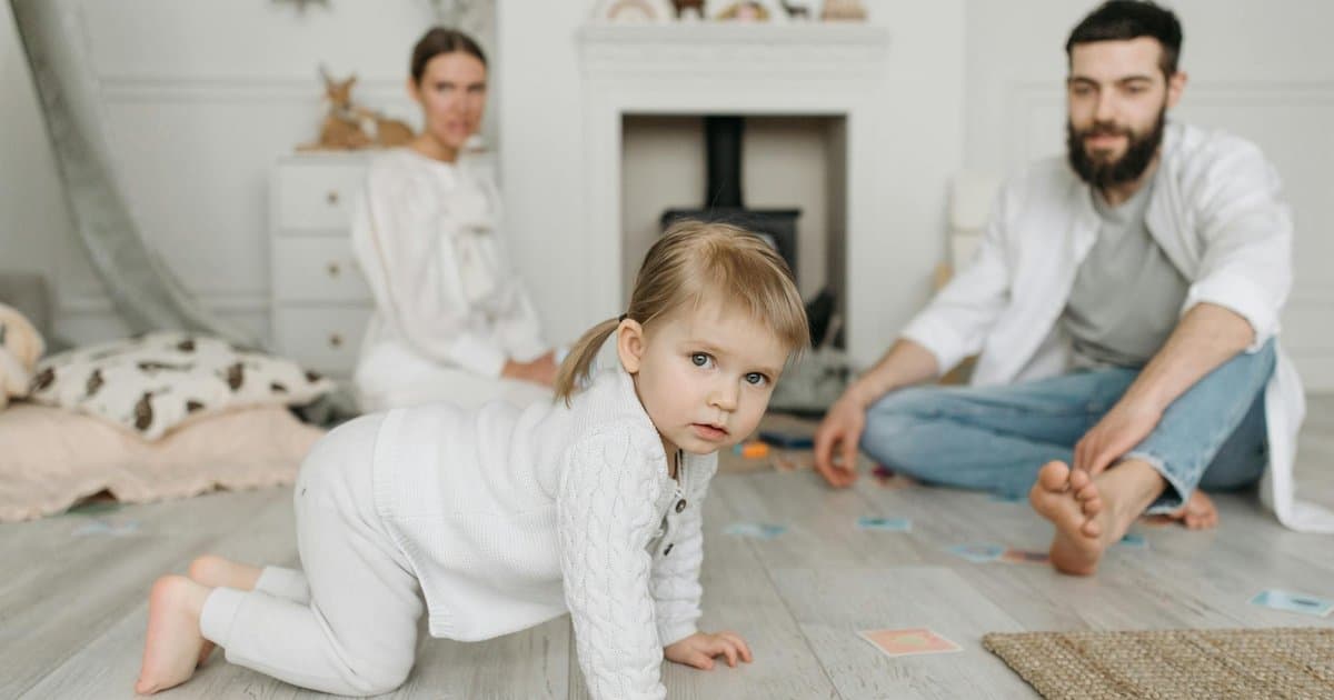Toddler in white sweater crawls on floor while parents watch in cozy room, illustrating meaningful first birthday gift ideas.