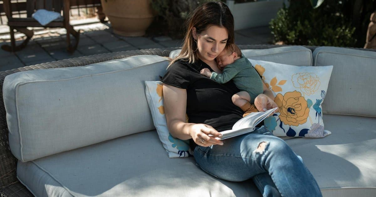 Mother reading to her sleeping newborn on an outdoor sofa, illustrating early bonding through books.